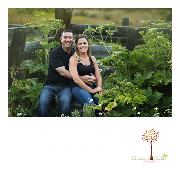 An engeged couple sit among green leafy plants and ranch fence while Sonora, CA engagement and wedding photographer Christine Dibble Photography takes their photos.