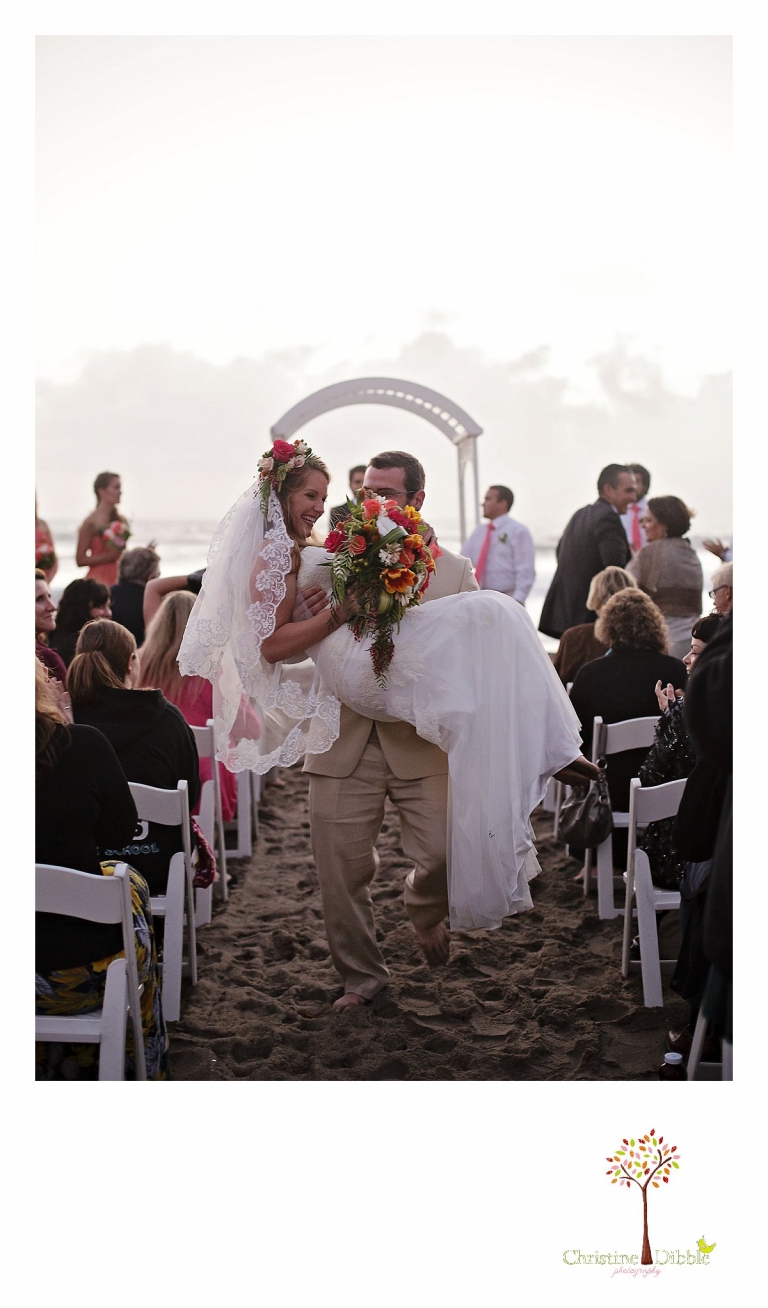 Sonora wedding photographer Christine Dibble Photography  photographs the groom carrying his barefoot bride down the aisle after the ceremony at their  sunset beach wedding at Moran Lake Beach in Santa Cruz.