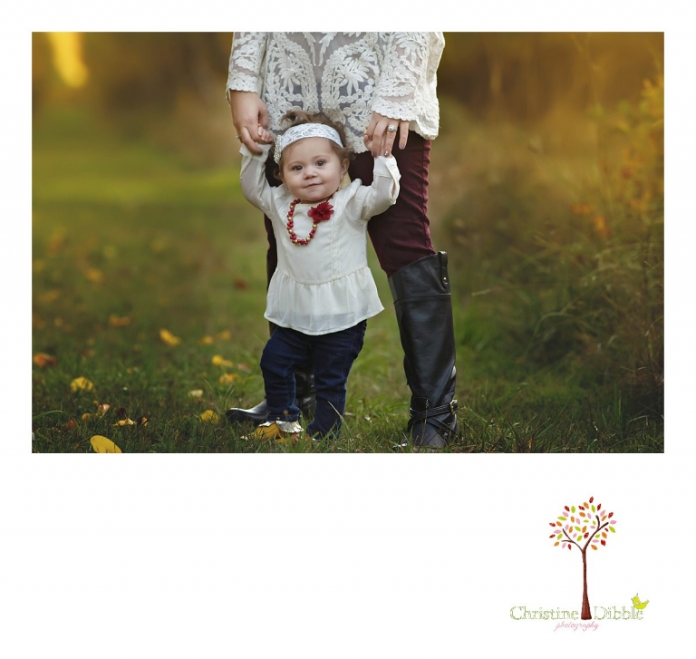 A baby girl hangs onto her mom's hands and stands on her boots during a Sonora family photography session photographed by Christine Dibble Photography at Indigeny Reserve in the fall among the apple orchards.