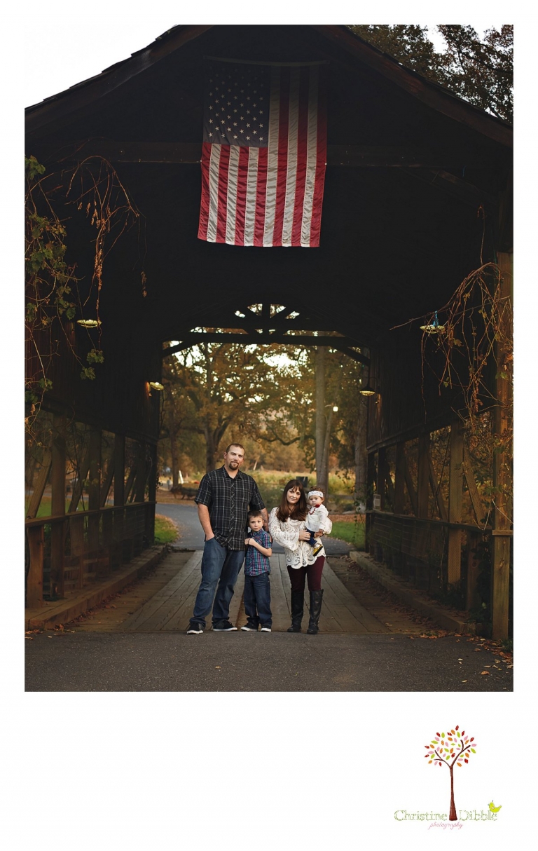 A family poses in front of the covered bridge during a Sonora family photography session photographed by Christine Dibble Photography at Indigeny Reserve in the fall among the apple orchards.