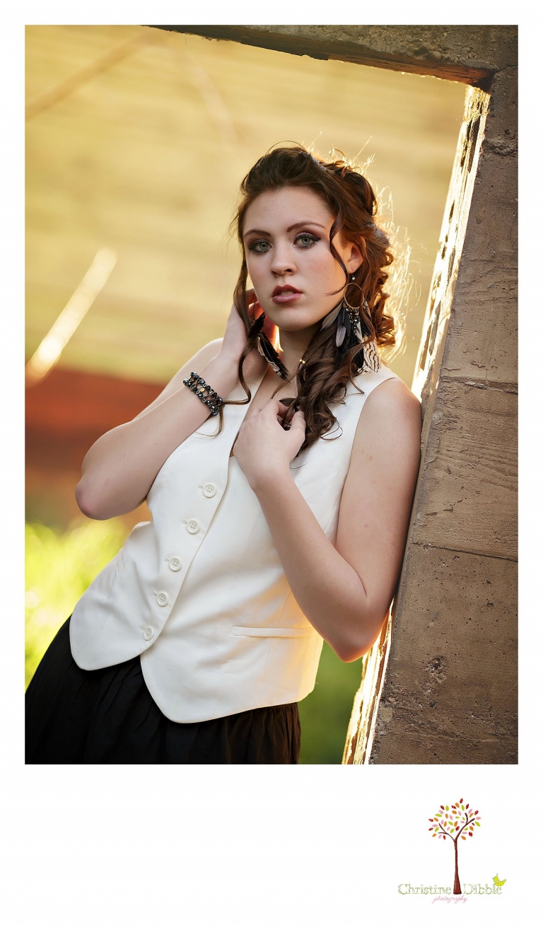 Sonora, CA senior model photographer Christine Dibble takes outdoor modeling photos of a teenage girl for a modeling portfolio while she wears dramatic make-up and feathered earrings and stands in a back lit door way.