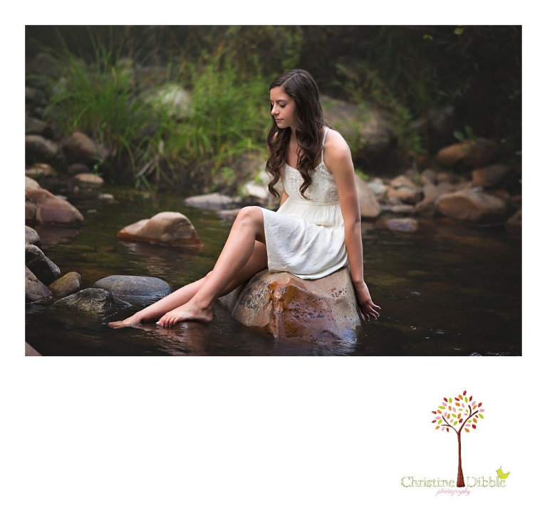 Best Sonora, Summerville and Turlock senior portrait photographer Christine Dibble Photography takes outdoor senior photos of a girl in a white dress dipping her fingers and toes into the river as she sits on a rock in the middle of the water.