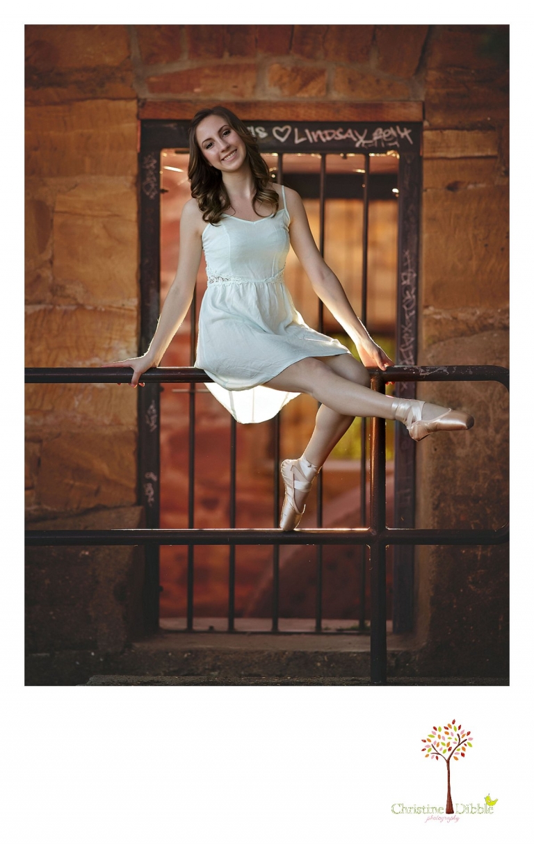 A dancer sits and poses on a bar in front of a window of backlighting while Summerville and Sonora High senior portrait photographer Christine Dibble Photography takes photos during a photography session.