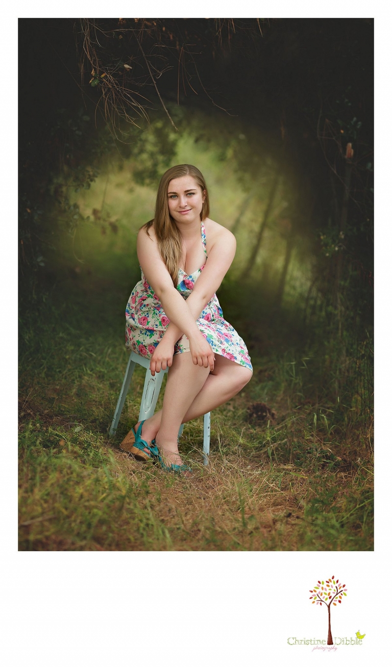 Sonora High senior portrait photographer Christine Dibble Photography takes rich color portraits of a senior girl in a floral dress sitting under a tree tunnel that is backlit.