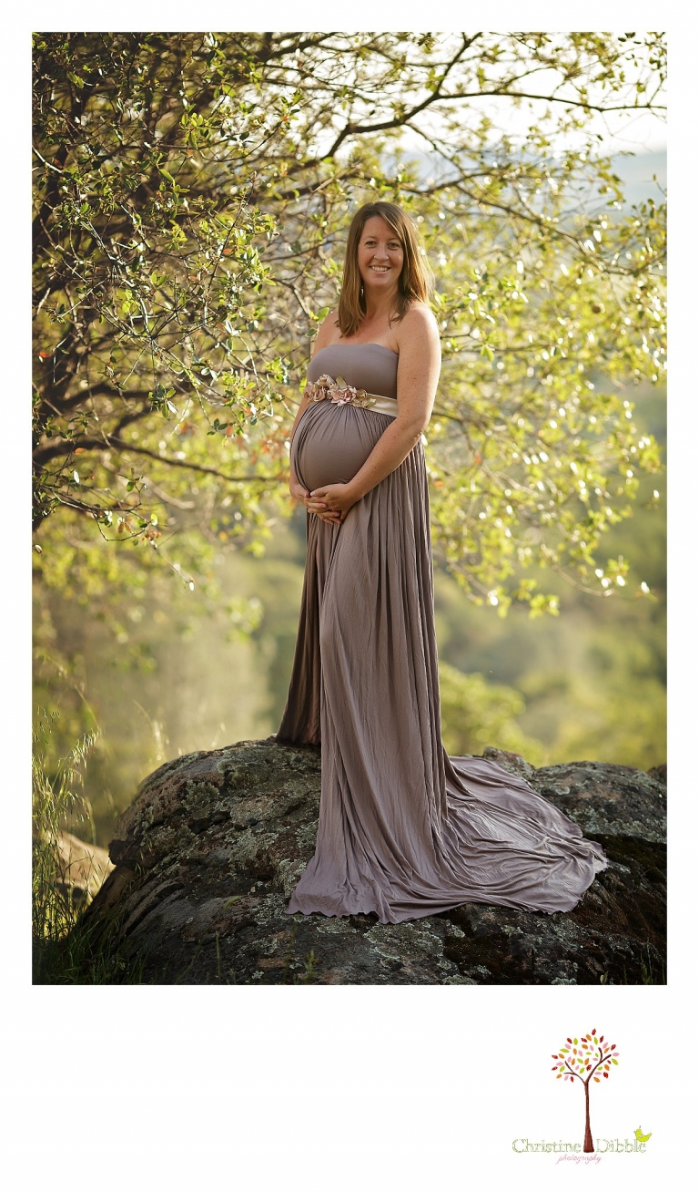 Sonora maternity photographer Christine Dibble Photography photographs a pregnant woman standing on a granite rock with a view behind her as she wears a maternity dress to show her belly which is decorated with a sash from Hopefully Romantic.