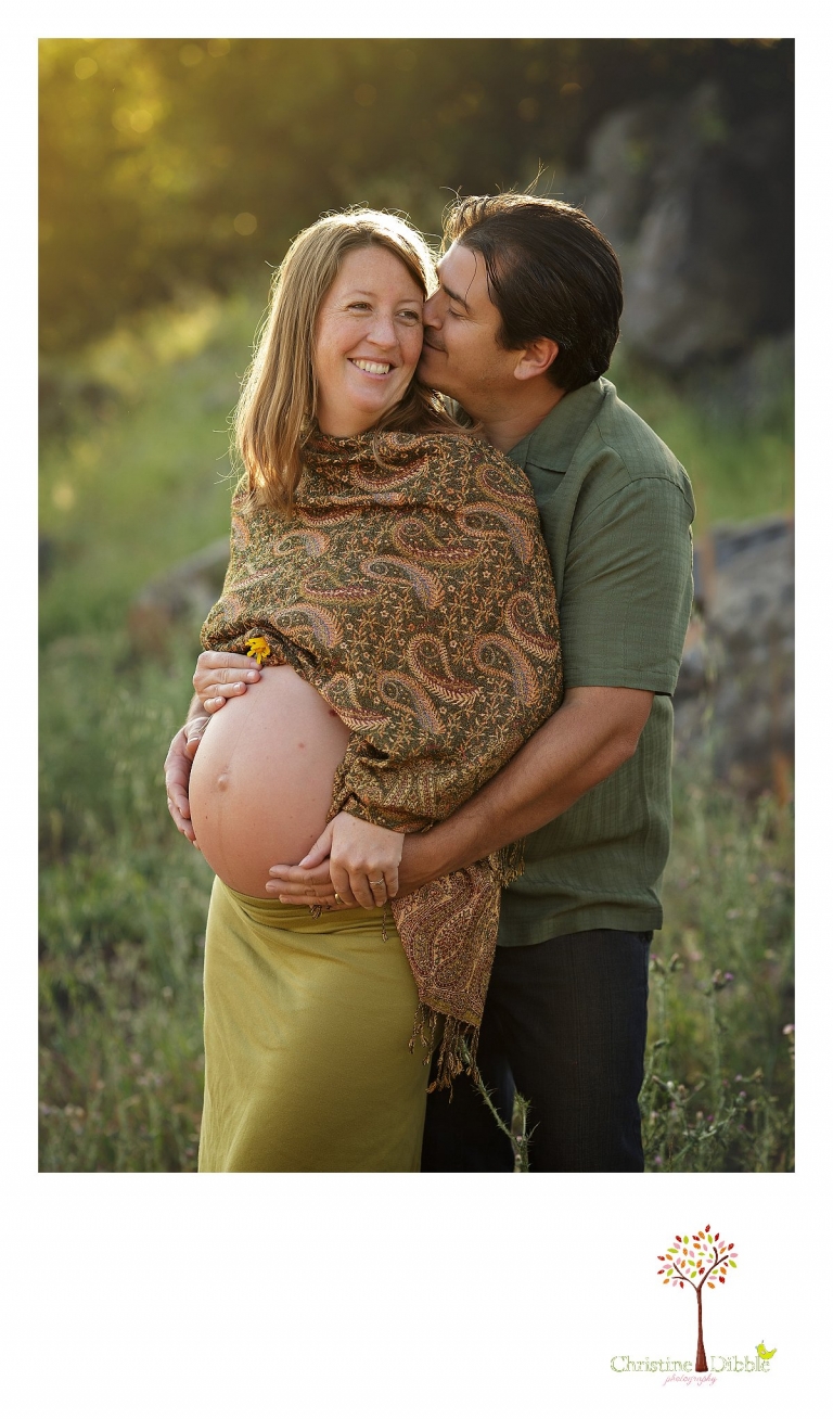 A dad-to-be kisses and whispers in mom's ear as they pose for Sonora maternity photographer Christine Dibble Photography during a sunset baby belly bump pregnancy photoshoot.