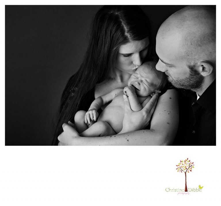 Sonora newborn photographer Christine Dibble Photography takes photos of a baby girl as her parents kiss her in a black and white portrait.