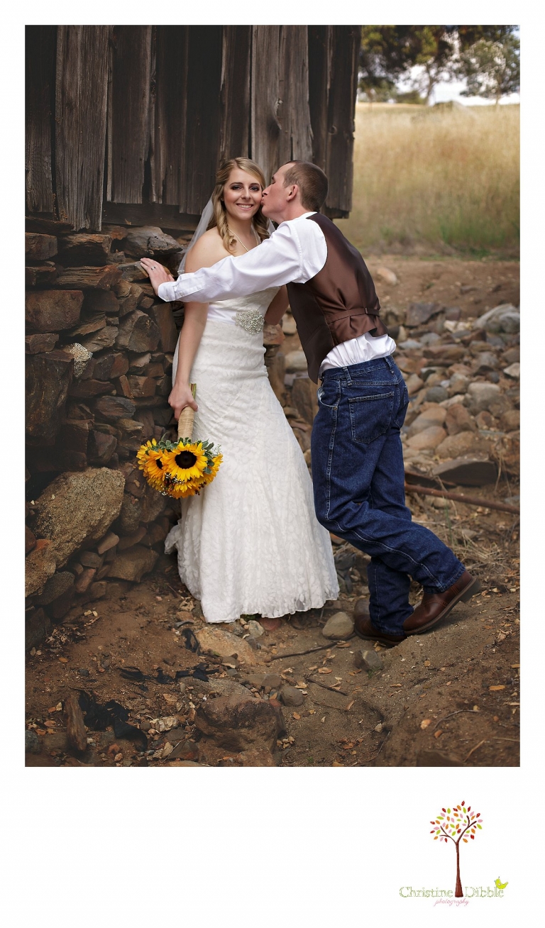 A groom kisses his bride on the cheek as they stand in front of a rustic ranch building at their Mother Lode Ranch wedding photographed by Sonora, CA wedding photograper Christine Dibble Photography.