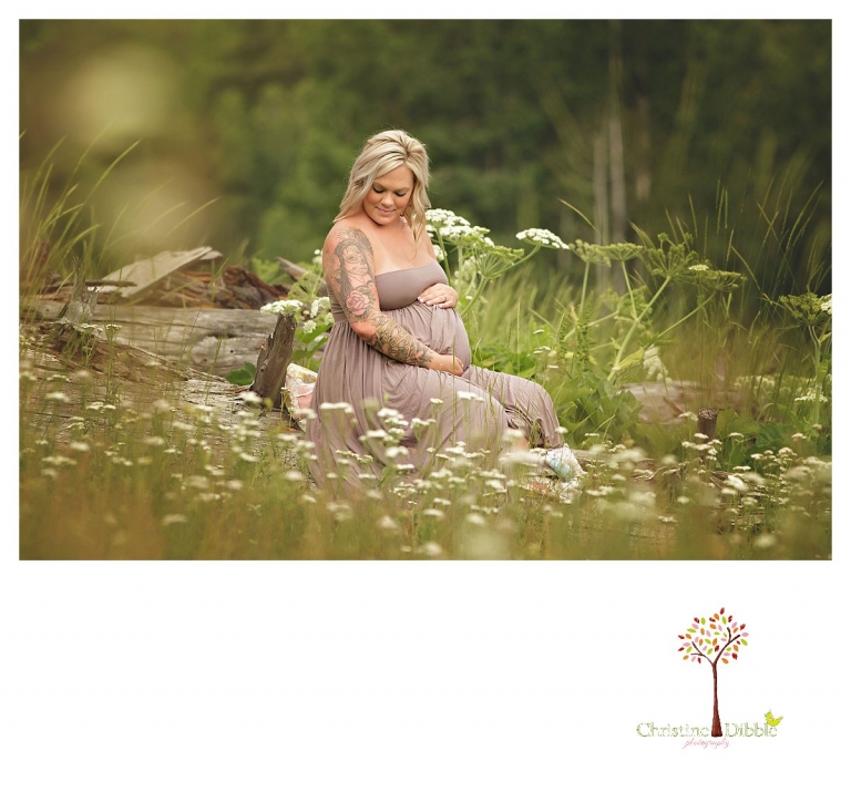 Sonora maternity photographer Christine Dibble Photography takes belly photos of a pregnant mother with tattoos sitting on a log in the woods among flowers.
