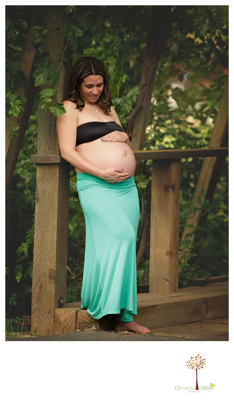 Sonora maternity photography session by Christine Dibble Photography shows a pregnant woman looking at her belly while she stands among grape vines under a covered bridge at Indigeny.