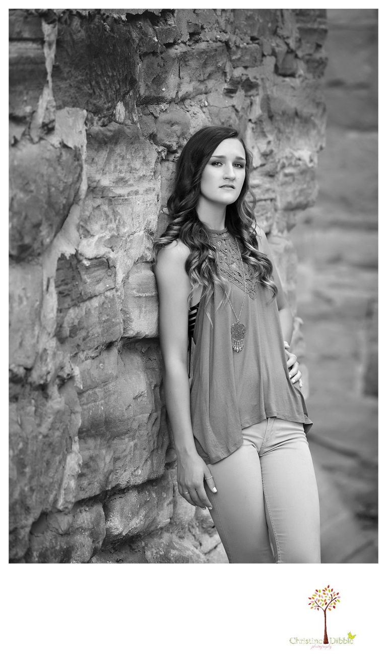 A senior girl leans on the wall of the mill at Knights Ferry while Summerville and Sonora senior portrait photographer Christine Dibble Photography takes black and white photos.