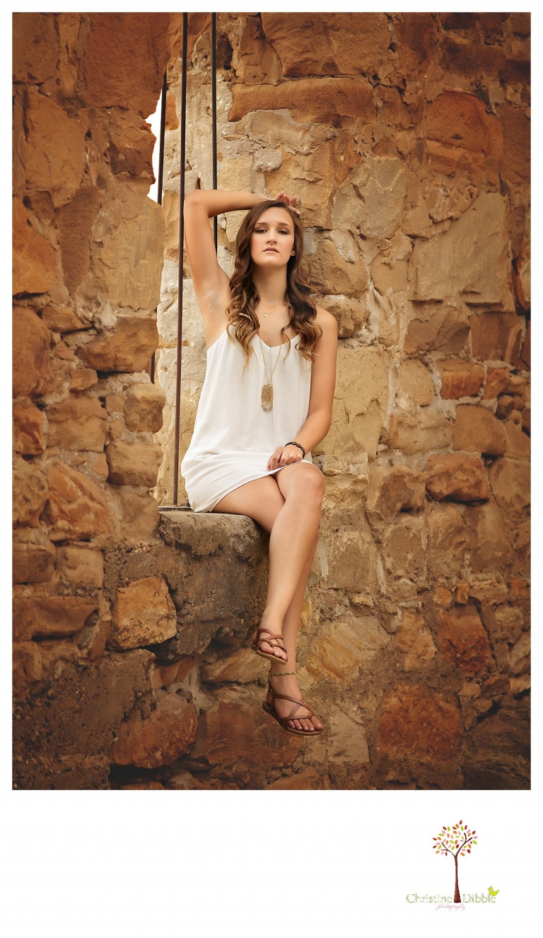 A senior girl in a white slip dress sits in the window of the mill ruins at Knights Ferry while Summerville and Sonora senior portrait photographer Christine Dibble Photography takes photos.