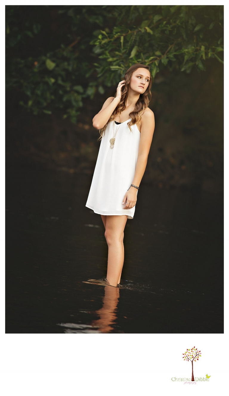 Standing in a white slip dress in the river, a senior girl is photographed looking over her shoulder by Summerville and Sonora senior portrait photographer Christine Dibble Photography.